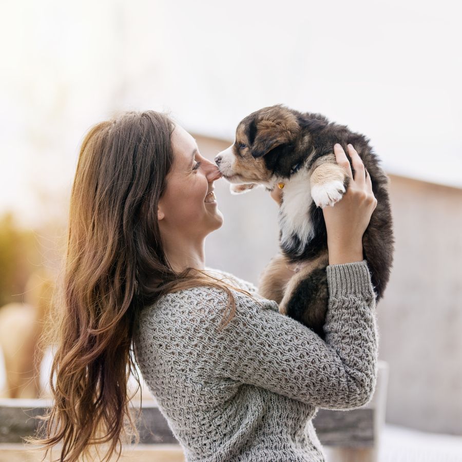 A woman in a gray sweater joyfully lifts a small puppy to her face, with their noses touching.